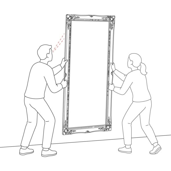 Two people carrying a large ornate-framed full-length mirror together — one on each side — approaching the wall to hang it, illustrating why a second person is essential for this step