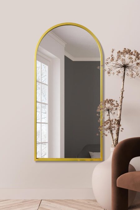 Tall arched gold mirror hung indoors beside dried botanicals and a velvet chair