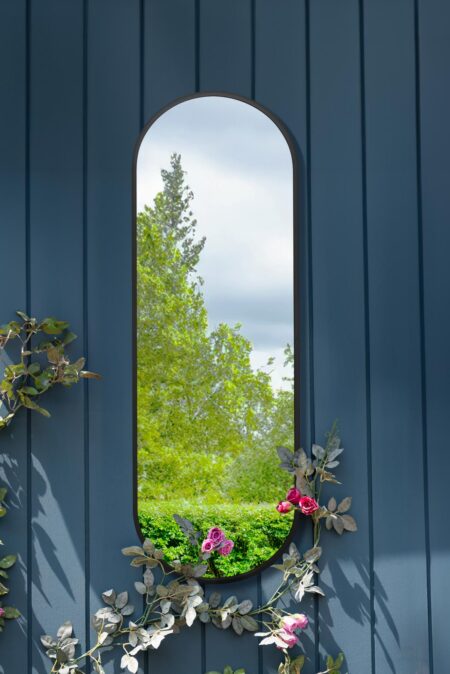 Black full length arch mirror with slim steel frame mounted on a blue garden wall with climbing roses