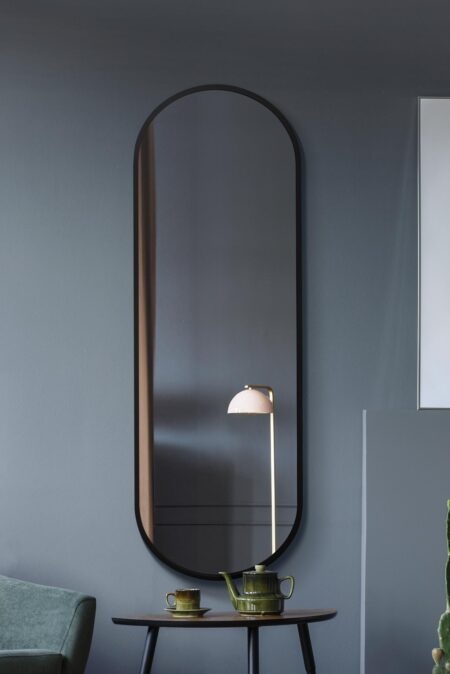 Tall capsule-shaped mirror with matte black frame styled above a wooden side table in a grey living room