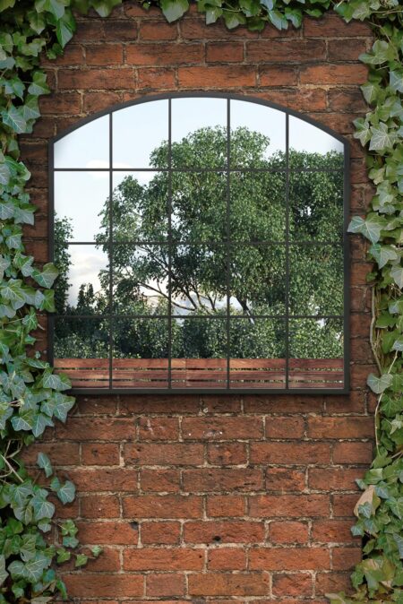 Black metal arch wall mirror with grid frame mounted on an ivy-covered brick garden wall