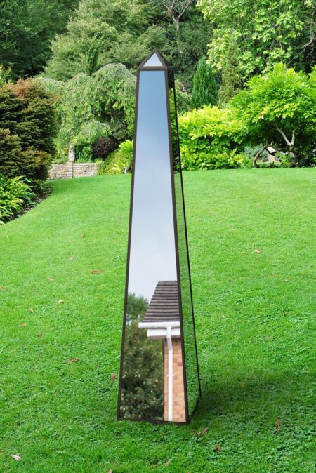 Tall mirrored metal garden obelisk with pyramid cap standing on a lawn surrounded by lush greenery