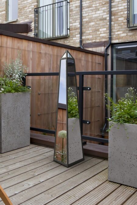 Mirrored obelisk garden ornament with black steel frame styled on a timber deck between concrete planters