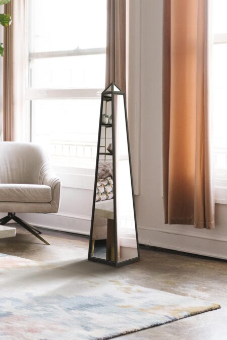Tall obelisk mirror with black metal frame standing beside a window in a light-filled living room