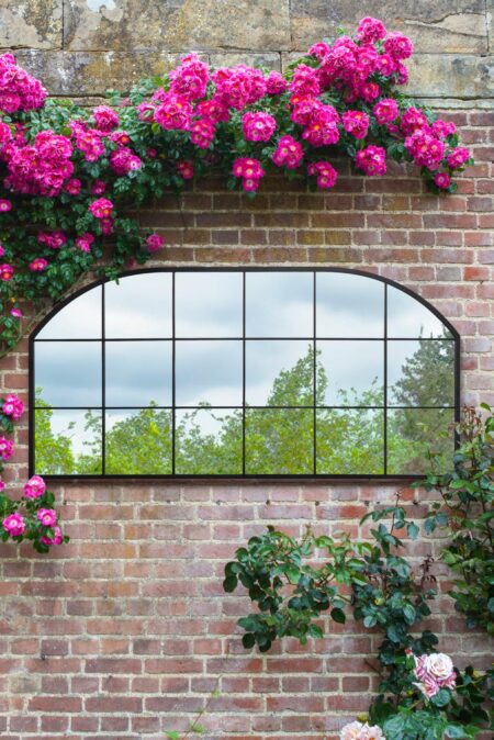 Black arch window mirror full length mounted on a brick wall surrounded by climbing pink roses
