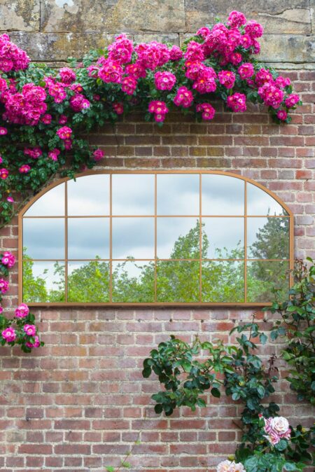 Gold full length wall mirror with arched windowpane frame mounted on a rose-covered brick garden wall