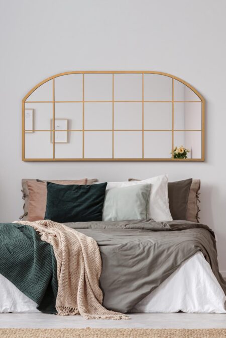 Wide arched gold windowpane mirror hung above a styled bed with layered neutral bedding