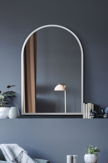 Arched white framed mirror displayed on a dark wall above a decorative shelf with houseplants and books