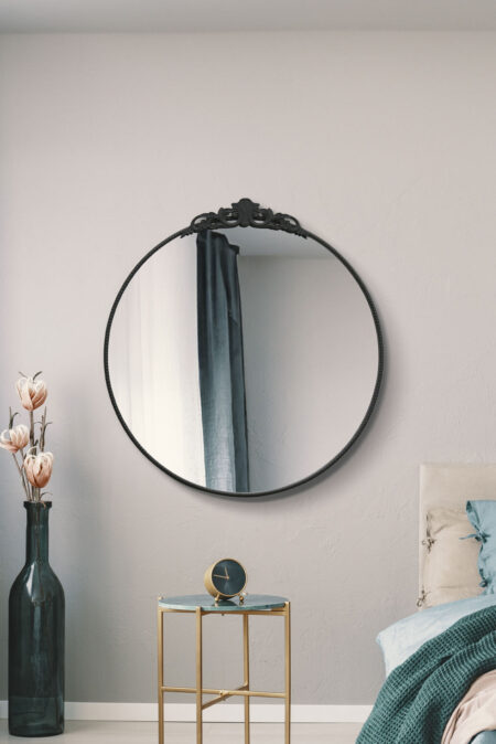 Large round black metal mirror with baroque crown detail hung in a styled bedroom