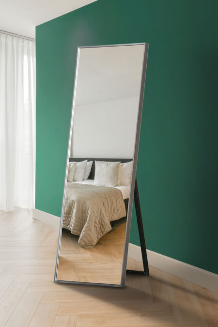 Silver full length mirror with stand leaning against a green bedroom wall