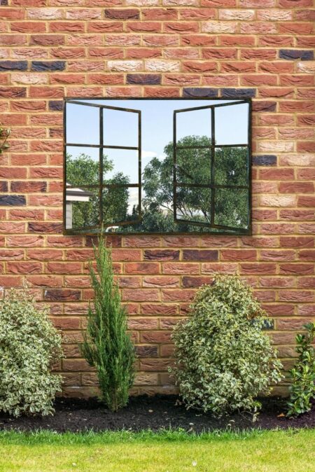 Black metal window pane mirror mounted on a red brick garden wall surrounded by shrubs